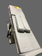 A medical examination table with padded white cushions, a metal frame, and an attached black control console, shown against a plain gray background.